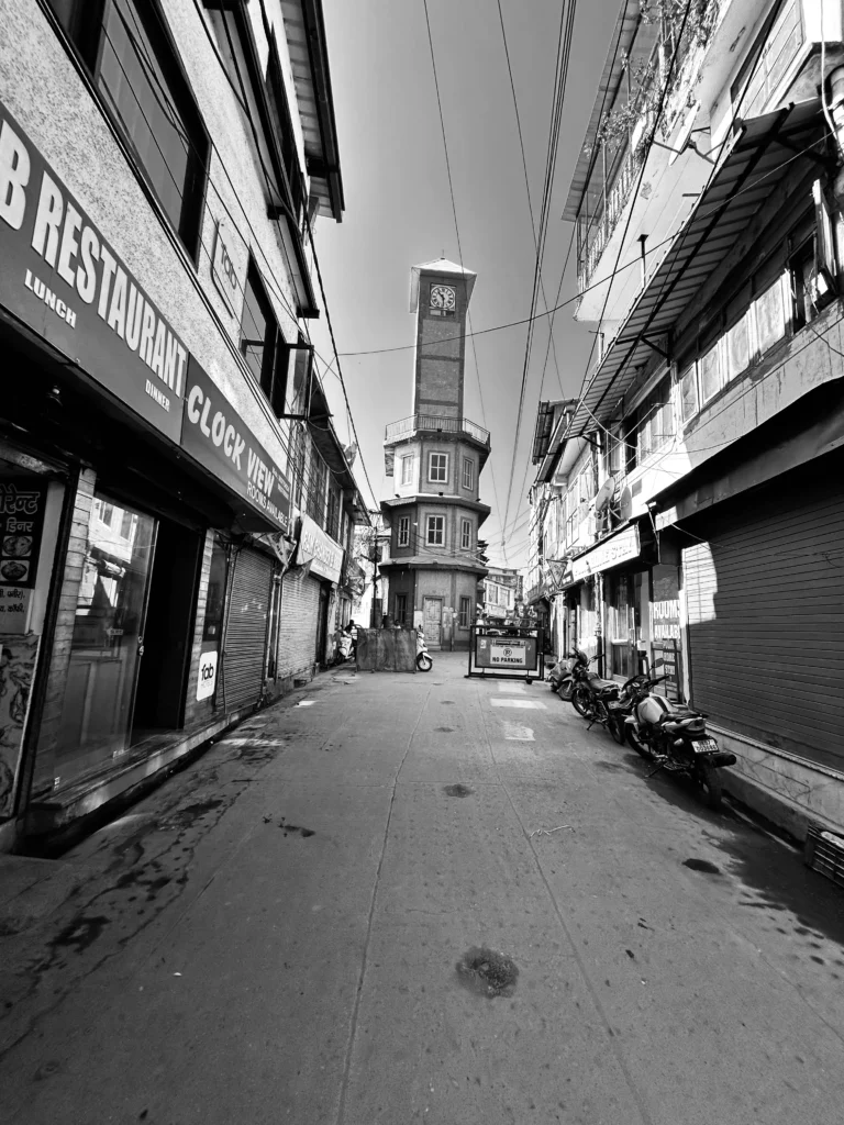 "Empty street with closed shops leading to a tall historic clock tower, captured in black and white photography."